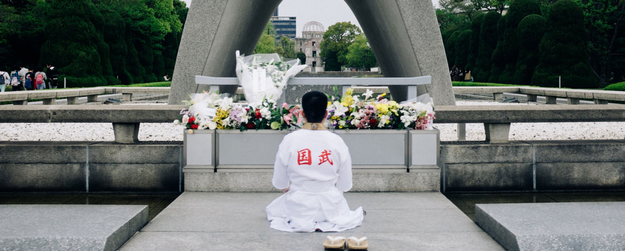 A man kneeling in front of the Hiroshima Memorial Cenotaph with flowers